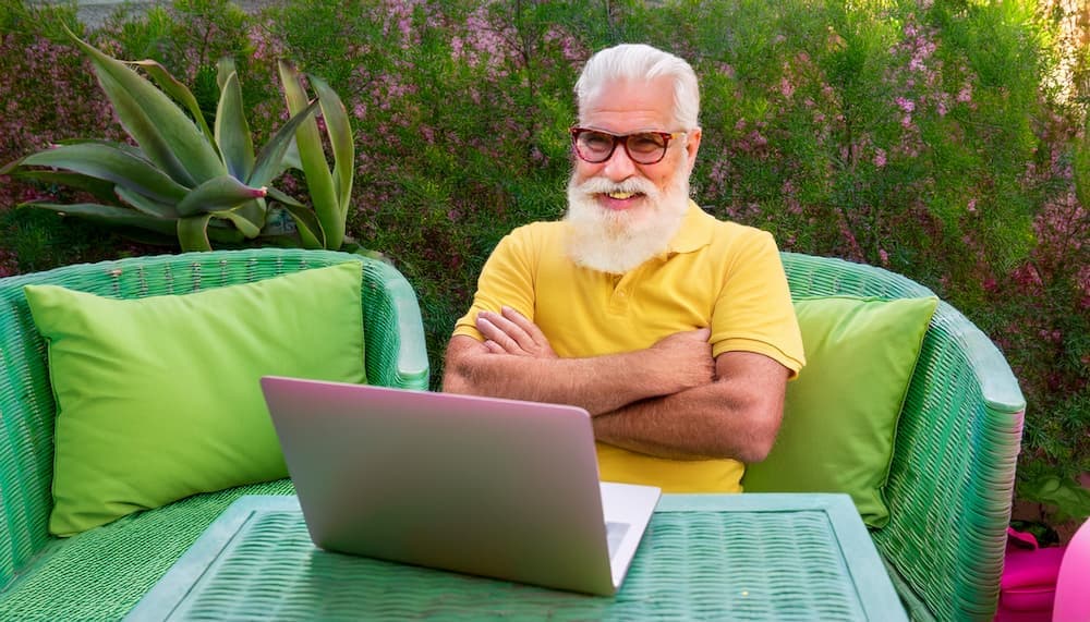Person sitting outdoors on green wicker furniture using a laptop on a table with plants in the background.
